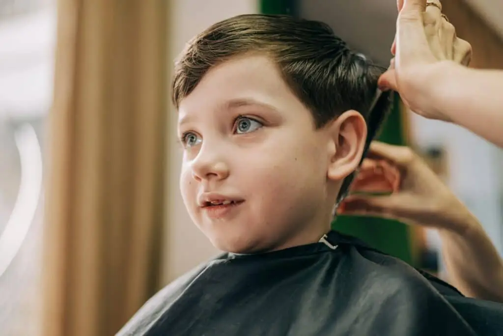 Little kid boy in the barbershop or hairdressing saloon. Barber is doing haircut for a toddler